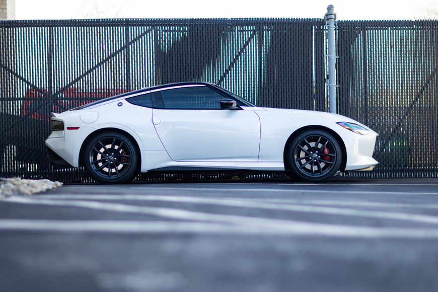 White 2023 Nissan Z parked in front of a chain-link fence, side view.