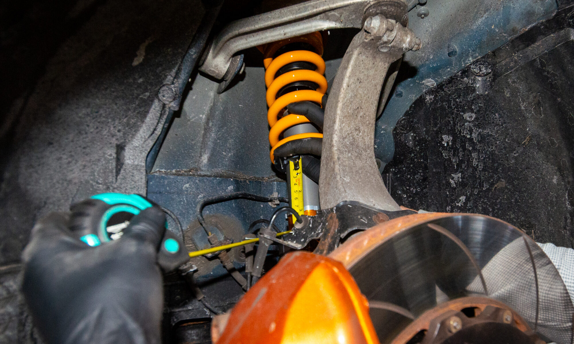 Mechanic measuring custom suspension with tape measure, showing orange coil spring and brake disc in a vehicle's wheel well.