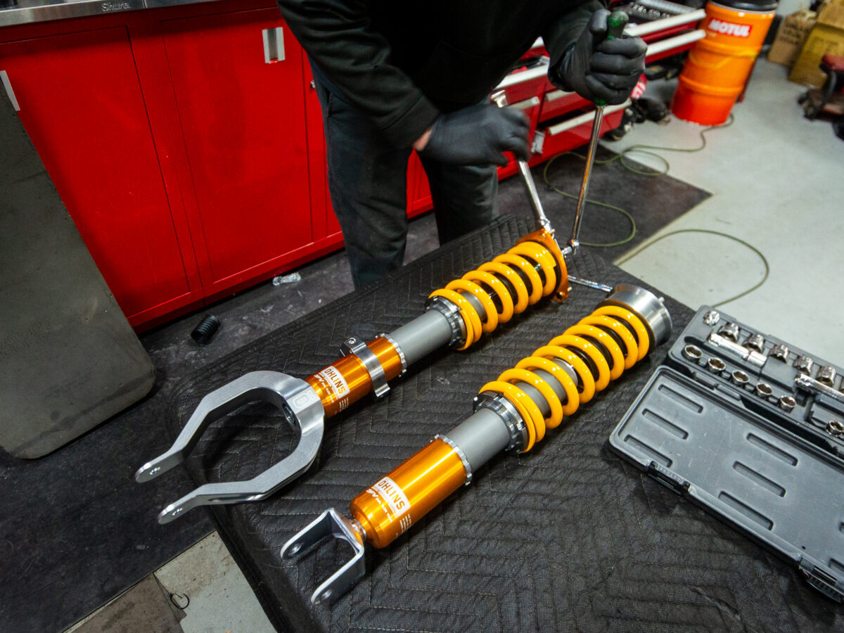 Mechanic installing orange coilover suspension on a workbench with tools nearby.