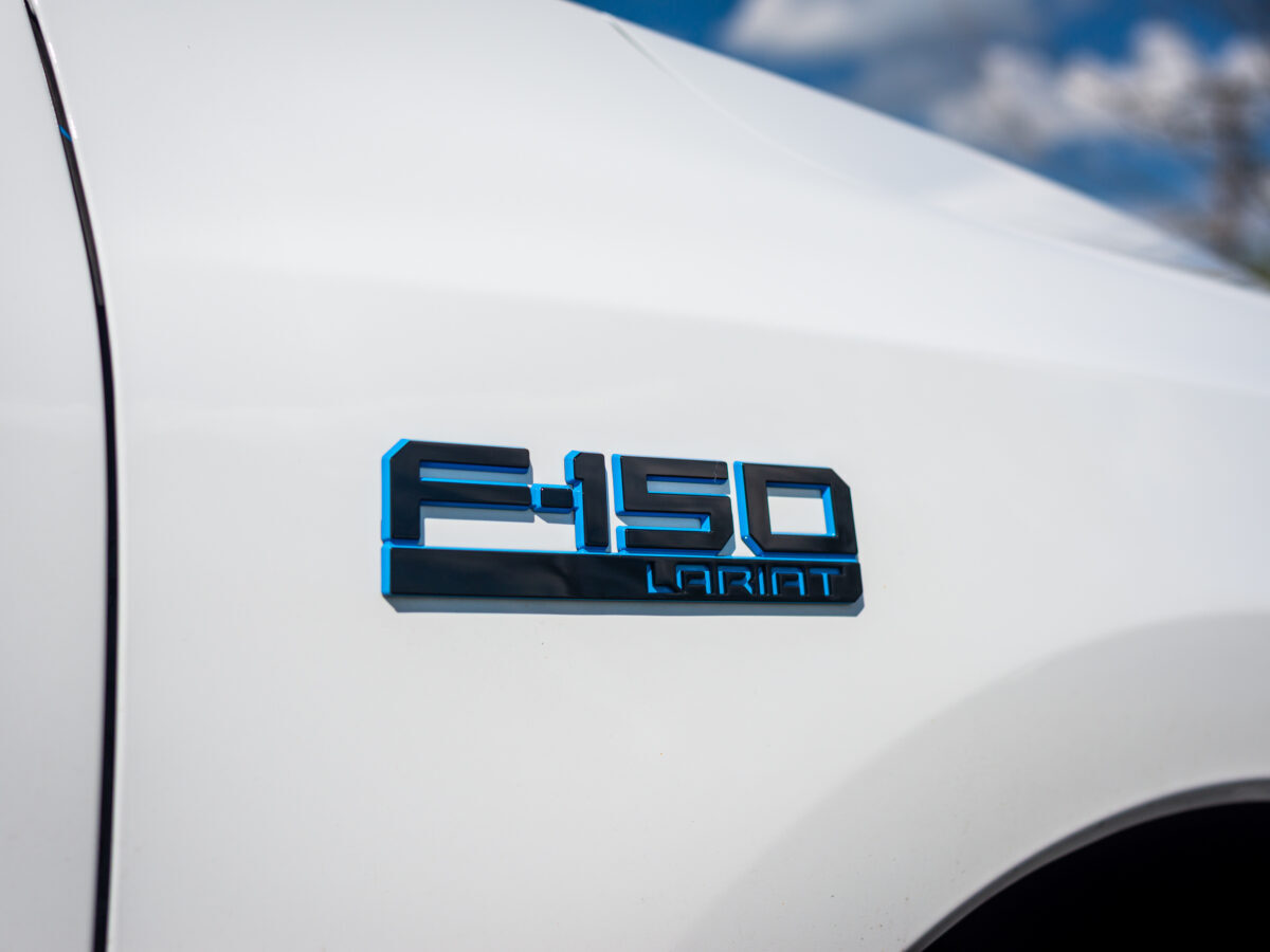 Close-up of a white truck's F-150 Lariat emblem with blue accents against a blurred sky background.