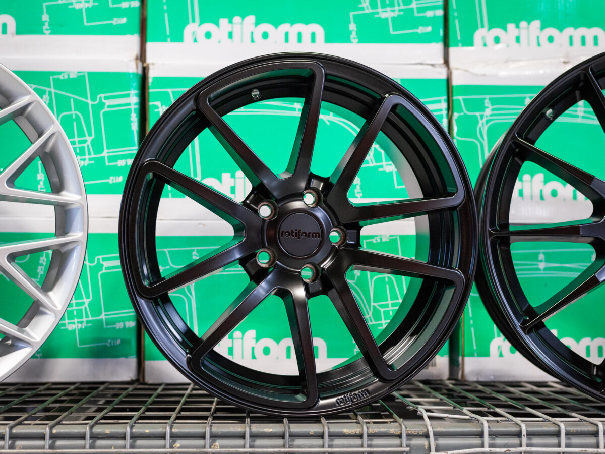 Black Rotiform wheel displayed between silver and another black wheel on a shelf with green boxes in the background.