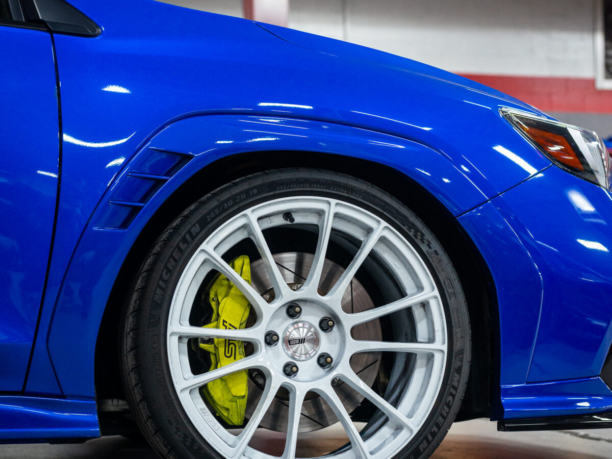 Blue Subaru with white alloy wheel and yellow brake caliper in a garage setting.