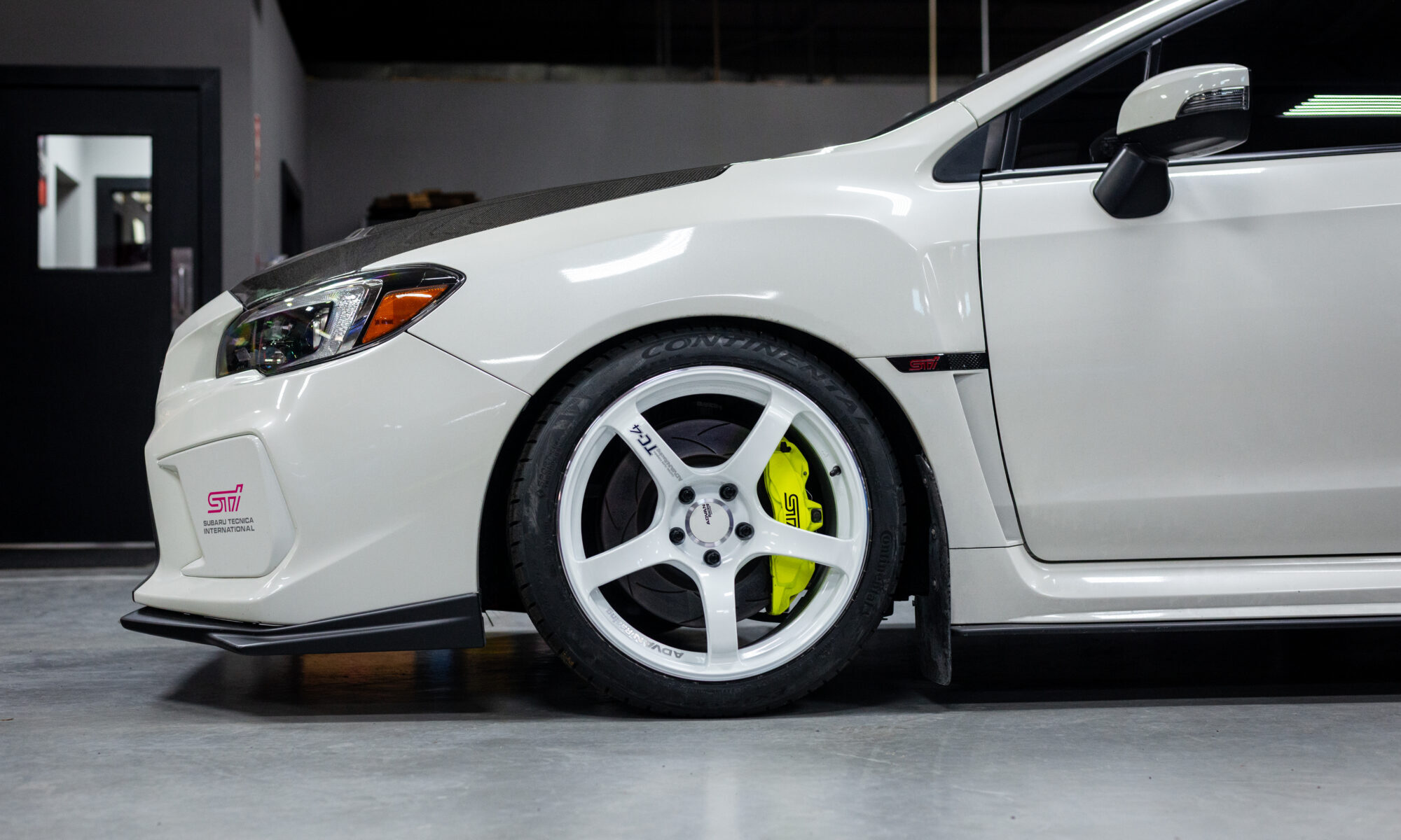 White Subaru with STI branding, featuring white wheels and yellow brake calipers in a garage setting.