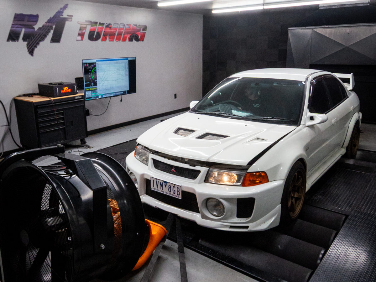 White car on a dynamometer in a tuning workshop, with a large fan and computer screen displaying data.