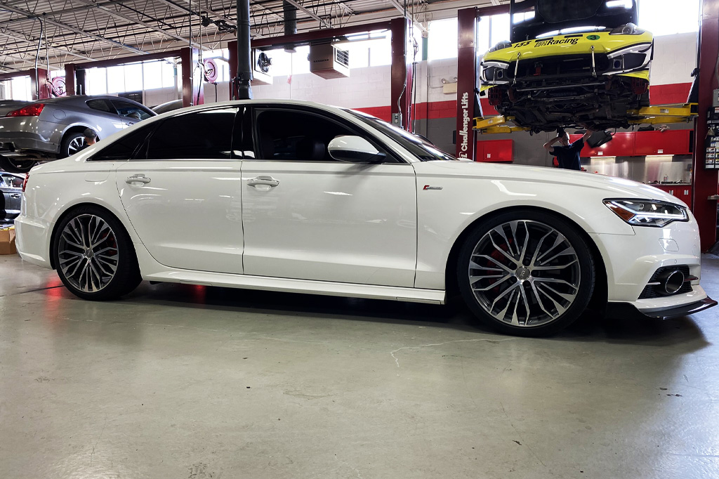 White sedan with tinted windows in a garage, lifted yellow car in background.