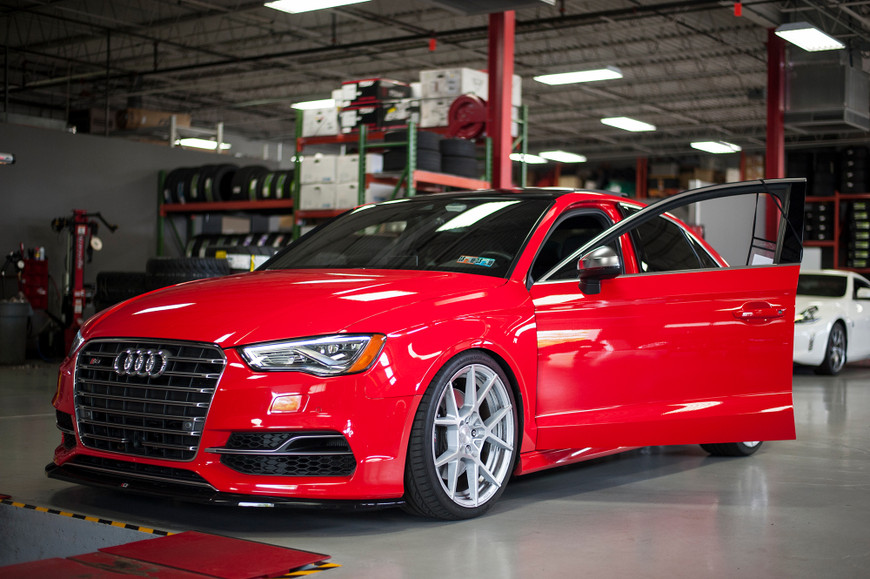 Red Audi S3 with open driver's door in a garage, surrounded by automotive equipment and tires.