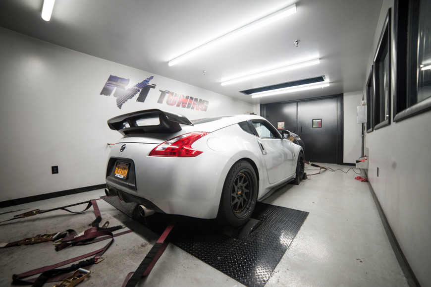 White Nissan 370z on a dyno in a tuning shop, rear view, with straps and equipment visible.