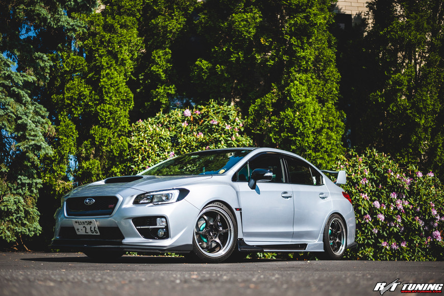 Silver Subaru STi parked in front of lush greenery, featuring Project Mu rotors and pads.