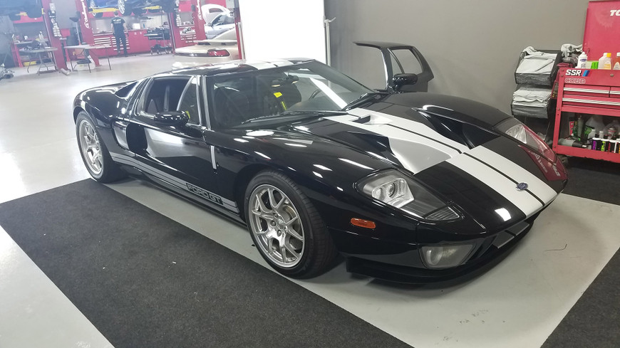 Black 2005 Ford GT with white racing stripes parked in a garage, driver's door open, red tool chest in background.